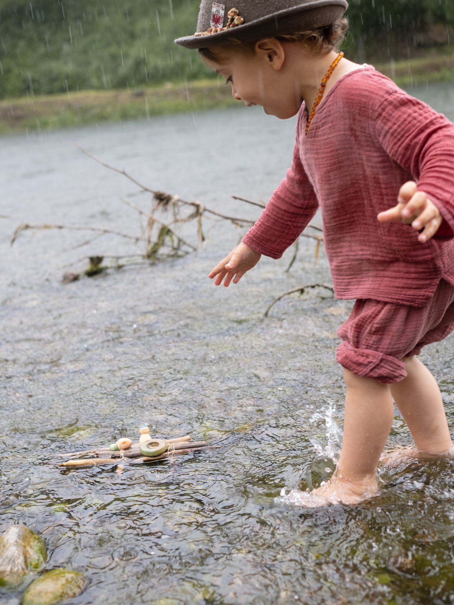 Child playing in a shallow stream wearing a red outfit and brown hat.