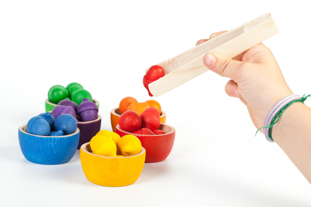 Using a pair of wooden tweezers to pick a colourful wooden acorn out of a small colourful wooden bowl