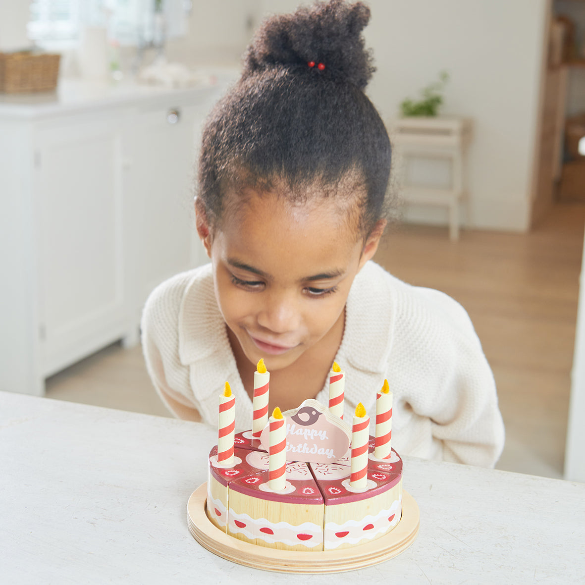 Child pretending to blow out candles on wooden toy chocolate birthday cake with six candles and cake topper which says happy birthday.  The cake is divided into six pieces and presented on a wooden plate.