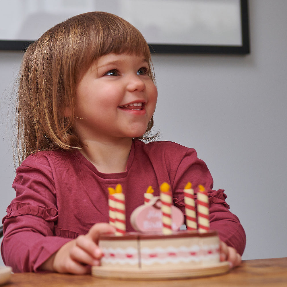 Child holding wooden toy chocolate birthday cake with six candles and cake topper which says happy birthday.